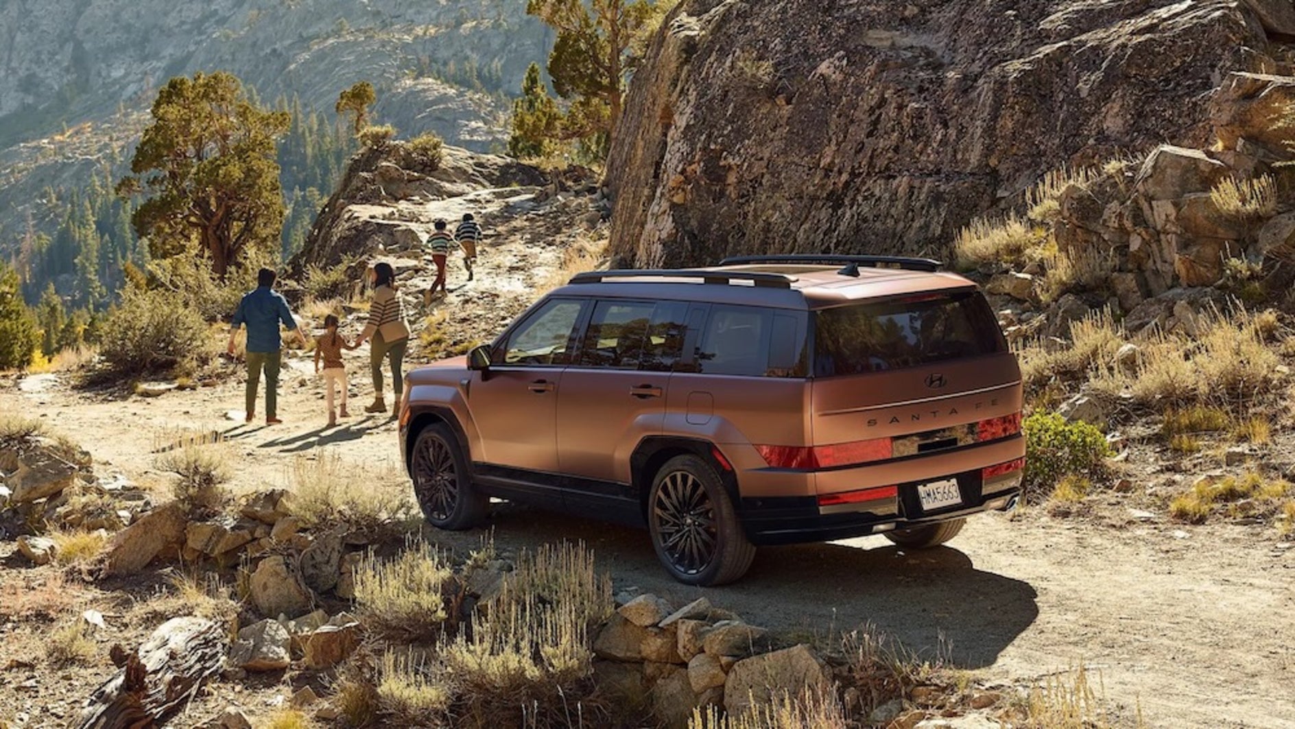 A family of four walking toward a mountain trail, with their copper-colored 2026 Hyundai Santa Fe SUV parked on a dirt road nearby
