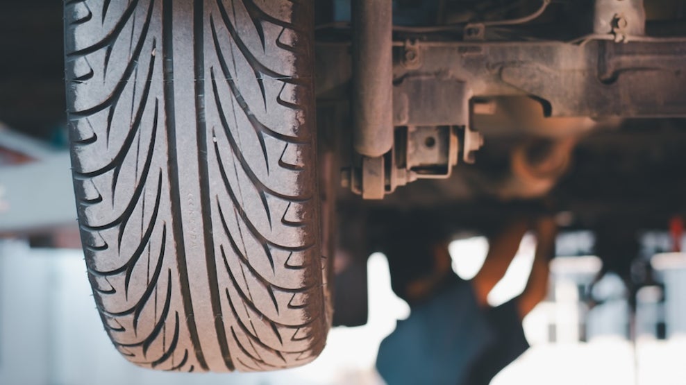 A closeup of a tire of a lifted car, with a mechanic working underneath the vehicle