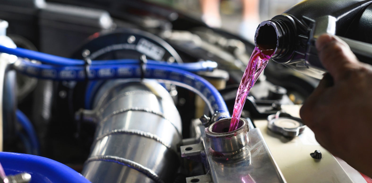 The hand of a person with brown skin pouring a bottle of pink antifreeze into an engine