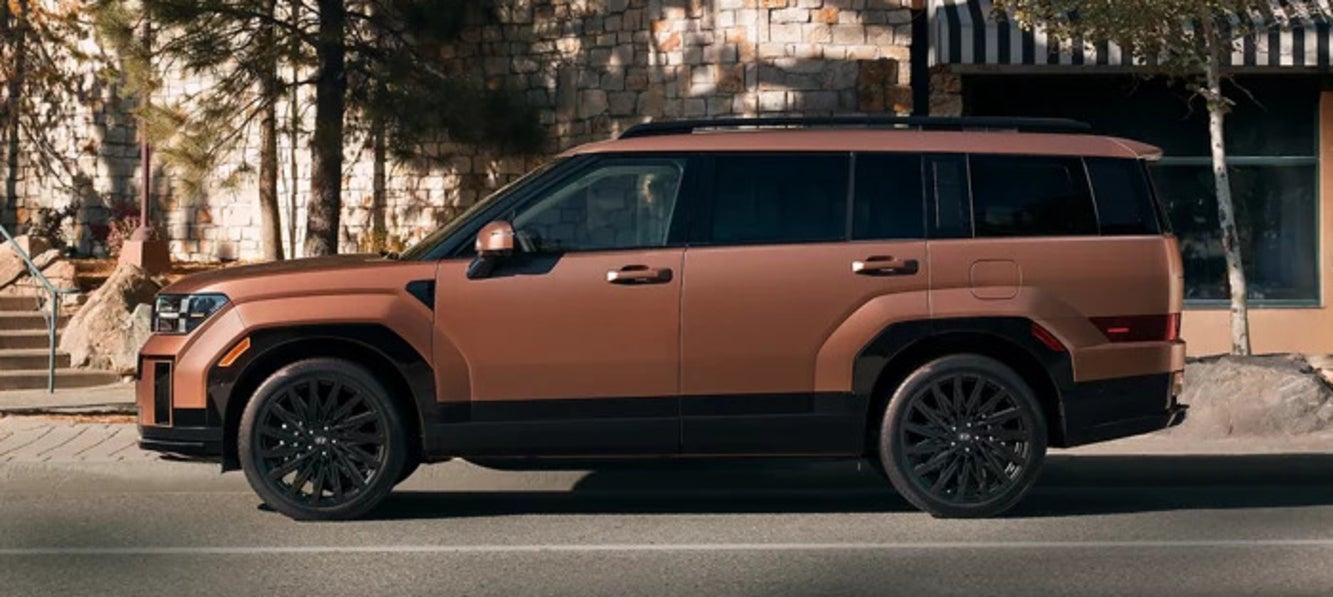 A side view of a copper 2025 Hyundai Santa Fe SUV driving past a stone building

