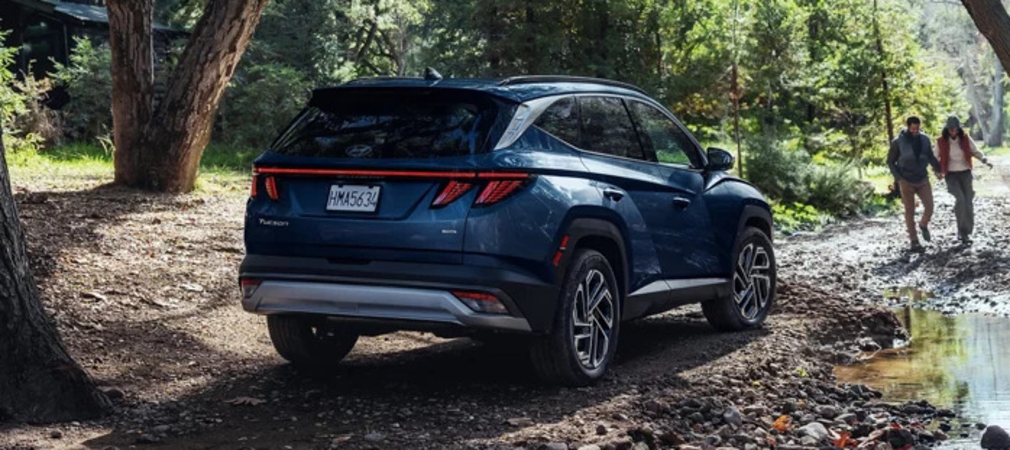 A dark blue 2025 Hyundai Tucson parked on a muddy forest trail, with two people talking towards it